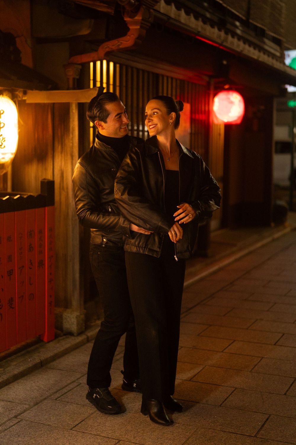 Couple in black leather jackets embracing and smiling on a lantern-lit Japanese street at night.