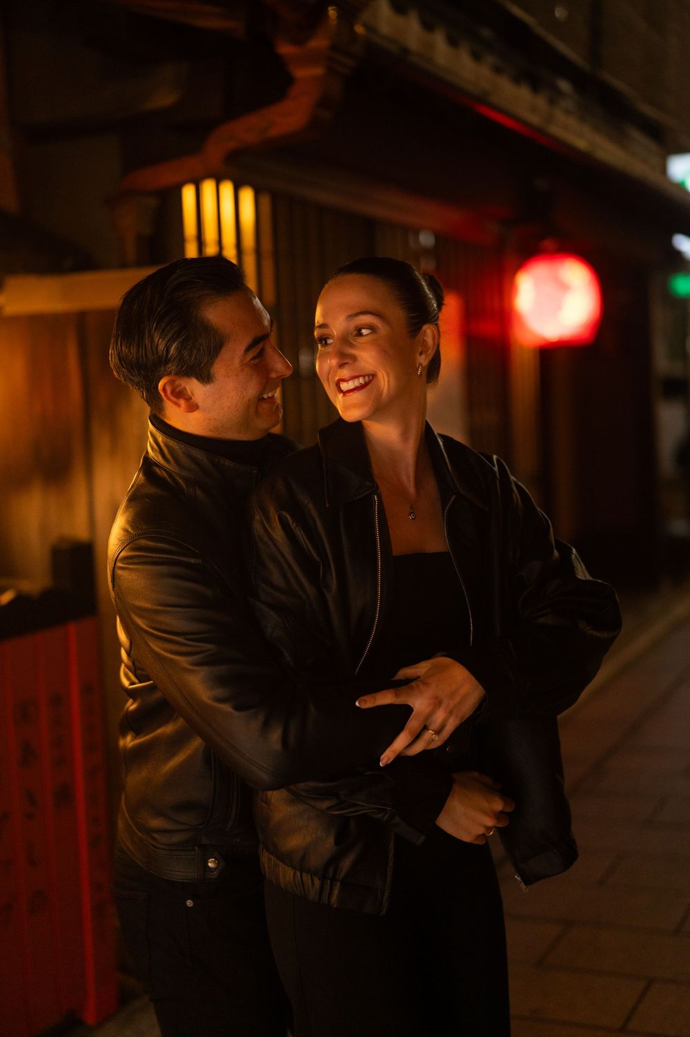 Couple in black leather jackets laughing and embracing on a warmly lit Japanese street at night.