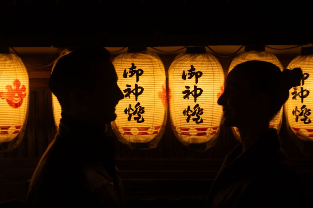 Silhouettes of a couple framed against glowing Japanese paper lanterns with kanji inscriptions at night.