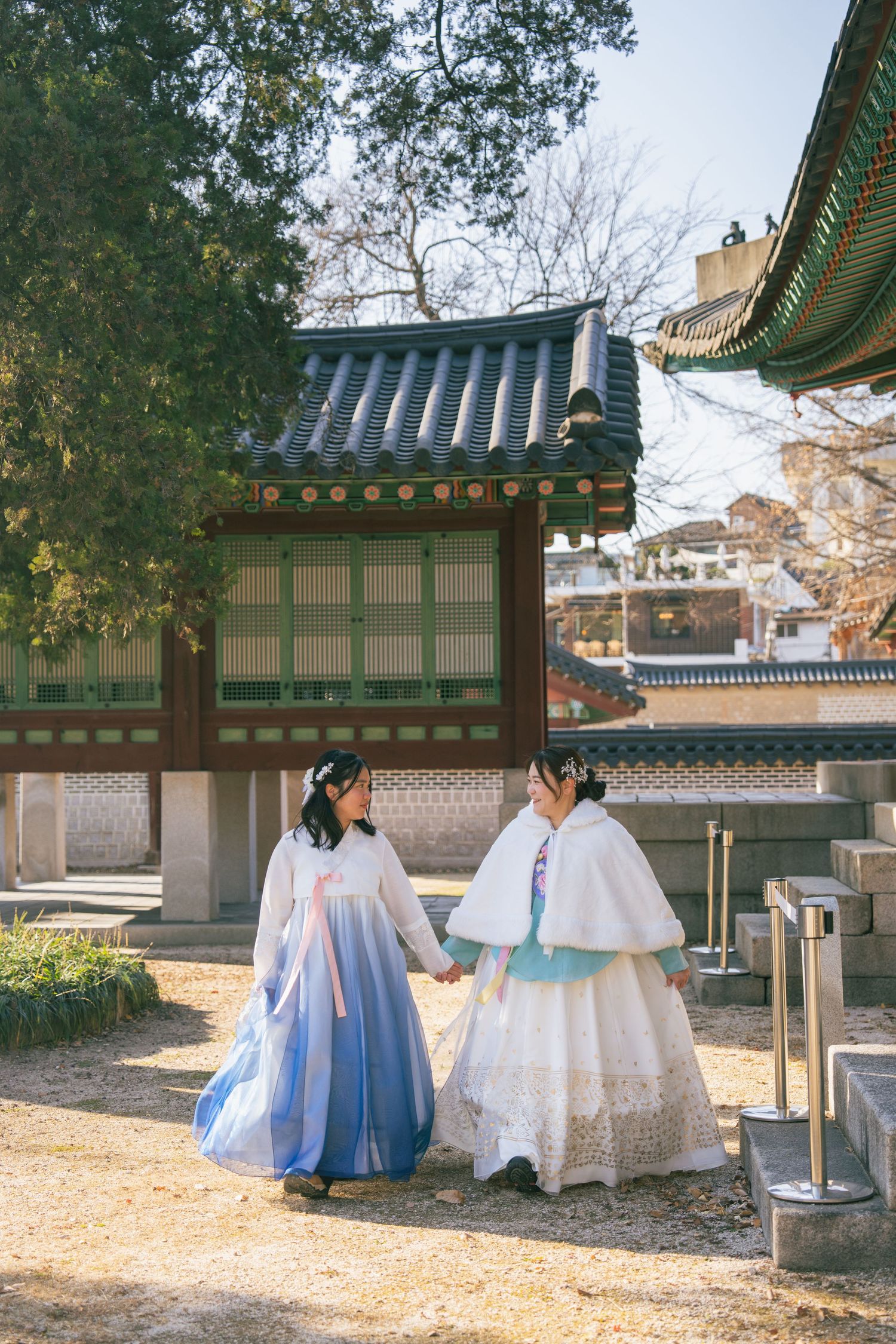 Two people wearing traditional Korean hanbok dresses sit and chat in a peaceful temple courtyard surrounded by autumn leaves.