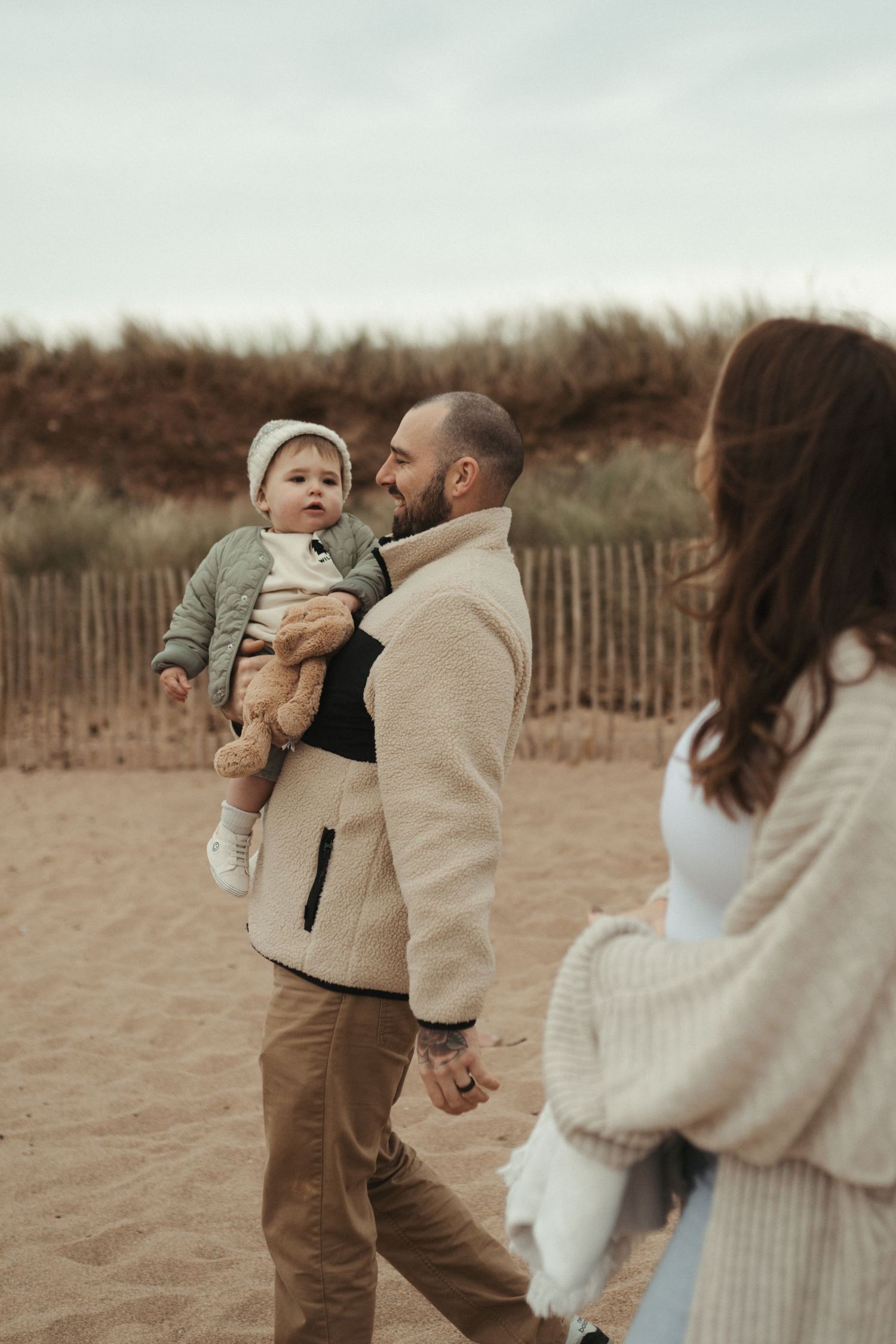 A family enjoying a playful moment together on a sandy beach with wooden fences in the background.