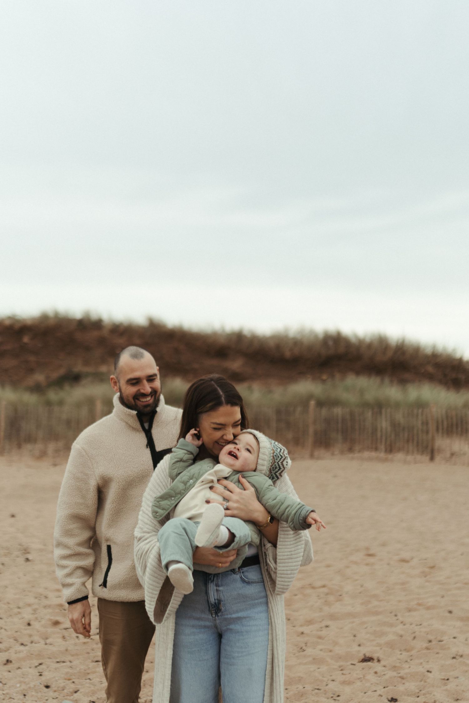 A family walks together on a sandy beach during an overcast day, wearing casual clothing.
