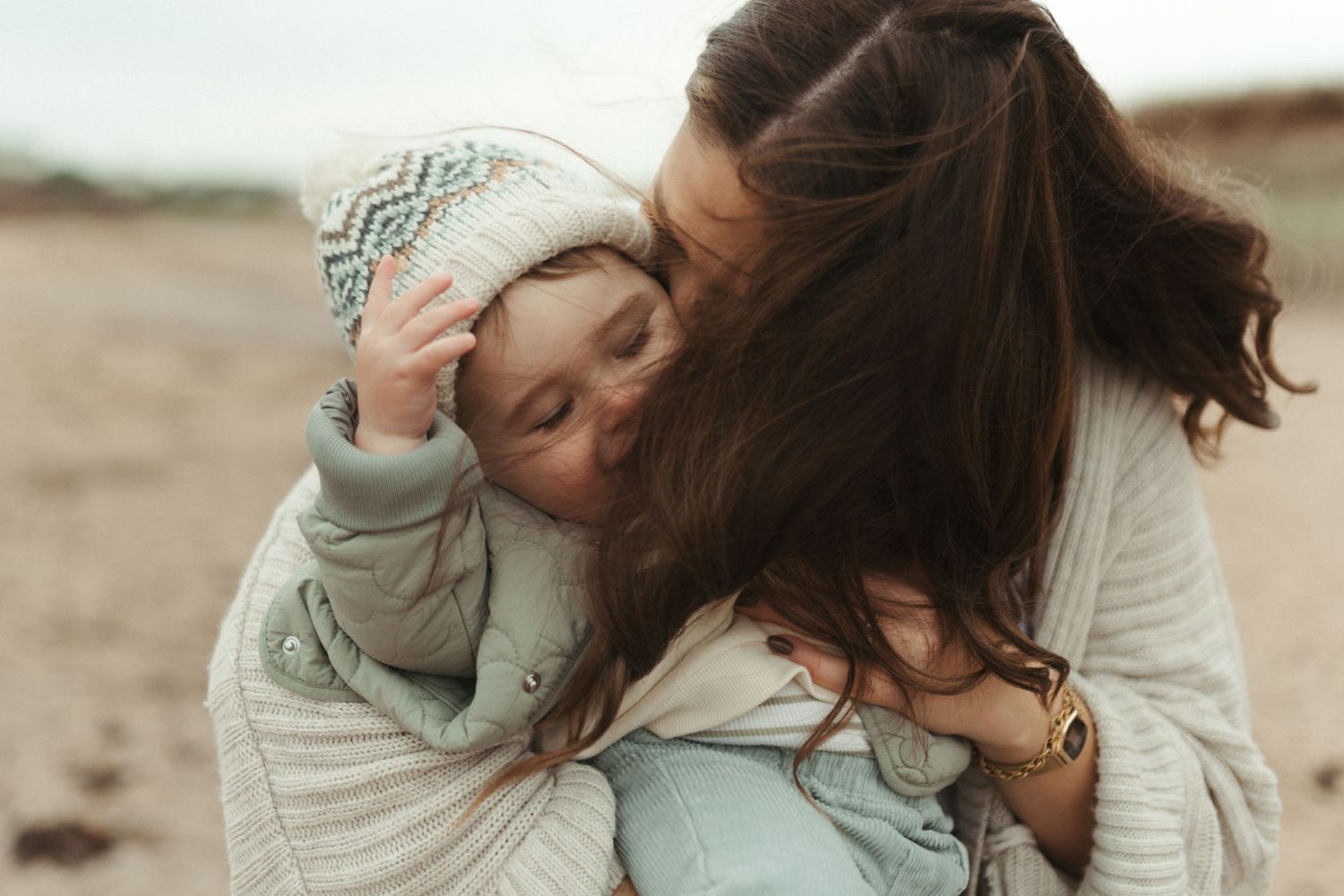 Romantic couple shares tender moments on a sandy beach wearing light colored sweaters and casual attire.