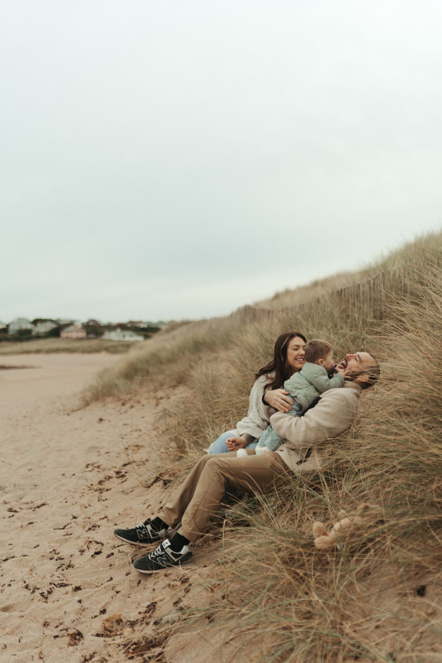 A couple relaxes together on a sandy beach dune with tall grass on an overcast day.