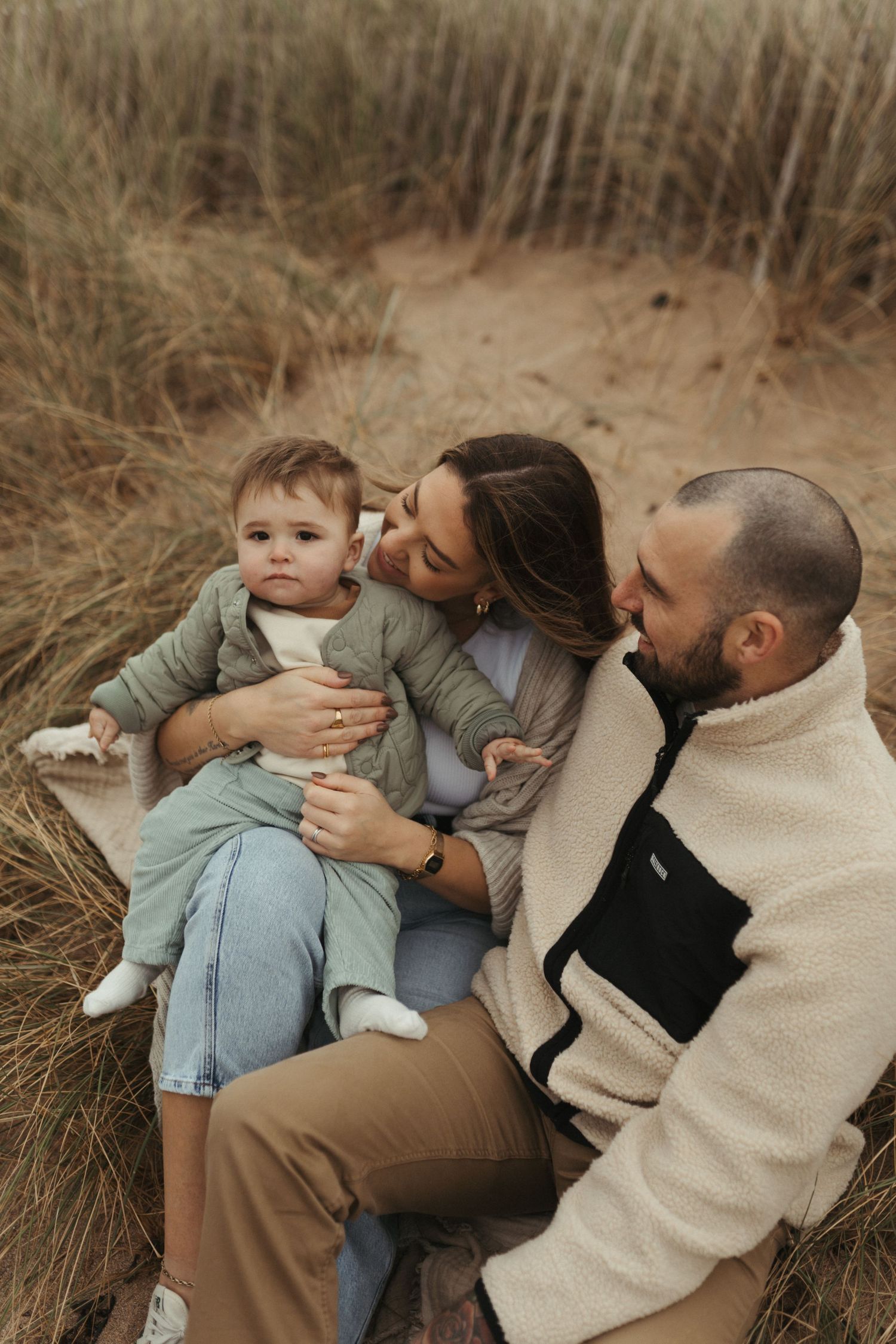 A family shares an intimate moment while sitting together among beach grass on sandy dunes.