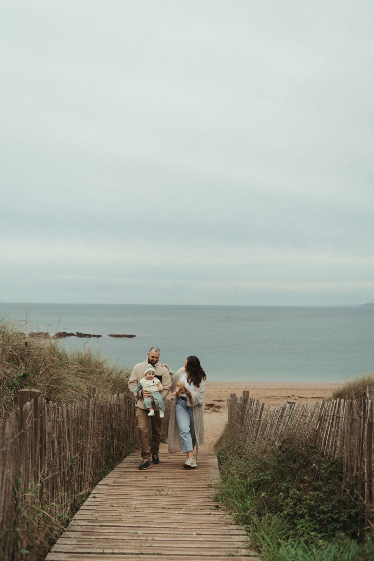 A wooden boardwalk leads to the ocean with rustic fencing and people walking in the distance.