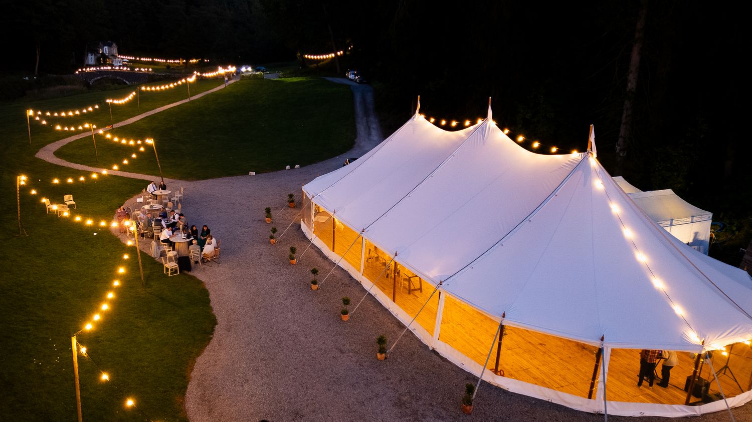 A white wedding tent illuminated by string lights glows warmly against the dark evening sky during an outdoor event.