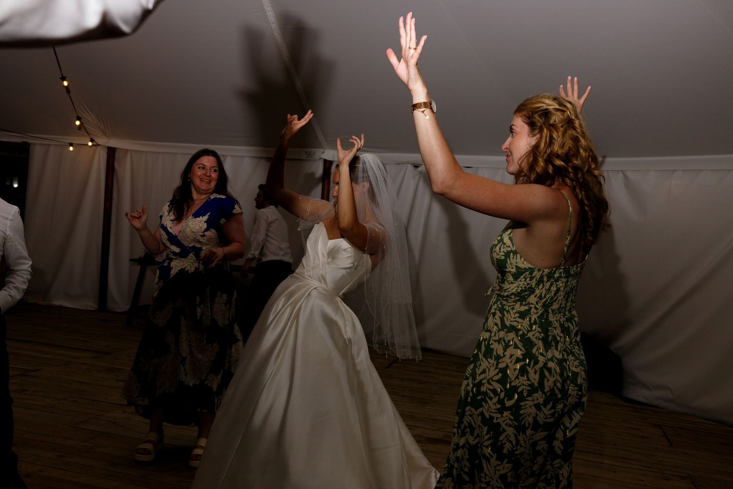 People dancing and celebrating at a wedding reception inside a tent with string lights overhead.