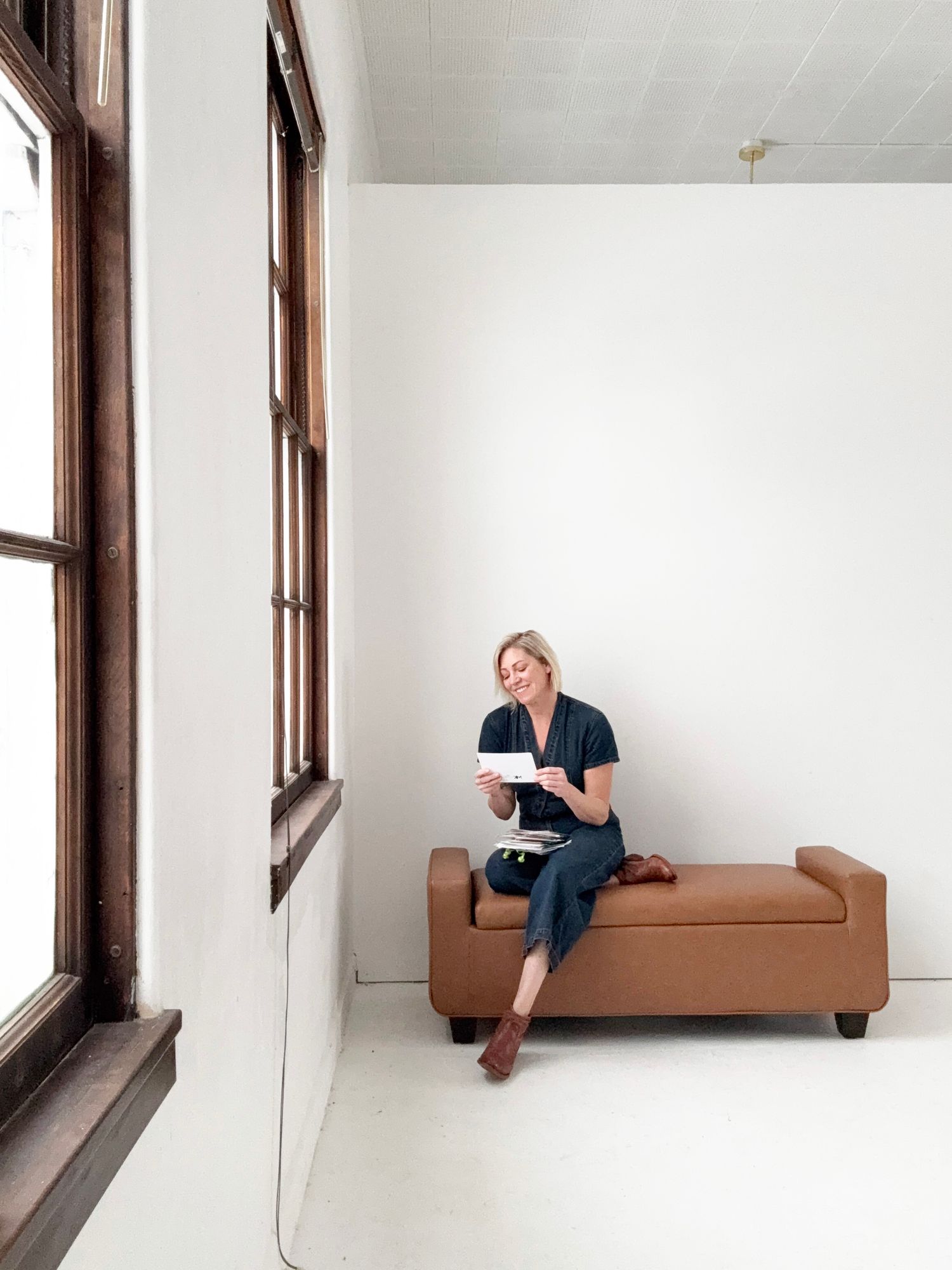 A person sits alone on a brown couch in a minimalist white room with large windows.