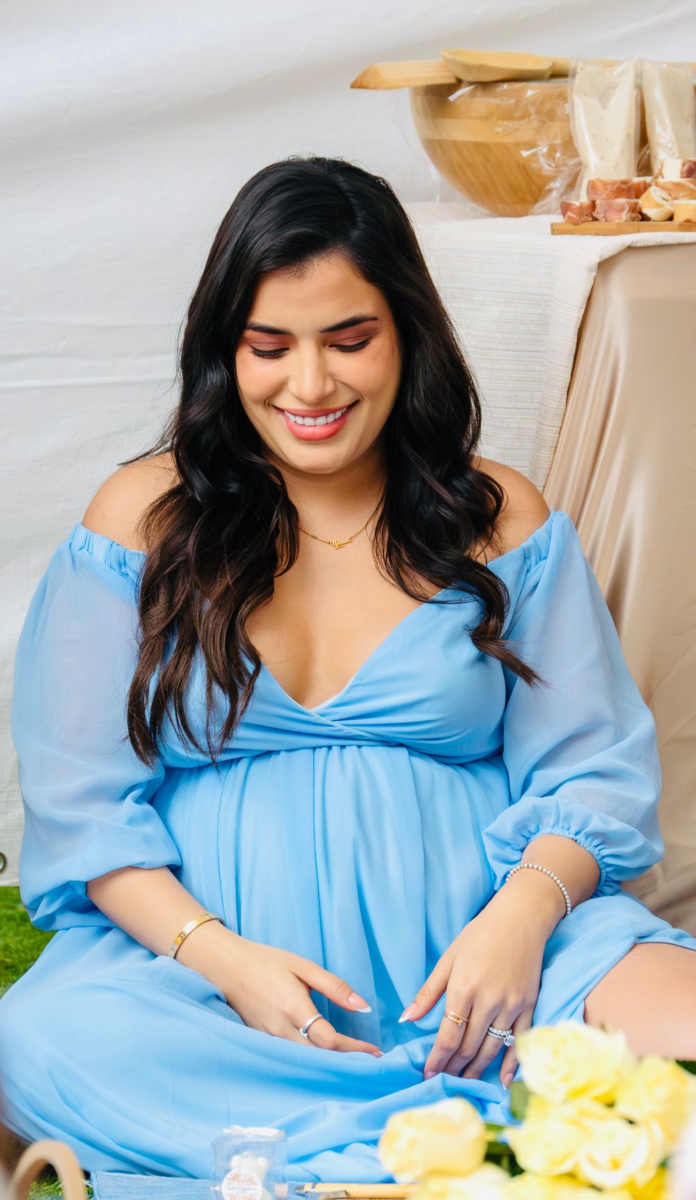 Pregnant woman in off-shoulder blue dress smiles while opening gifts at her baby shower celebration.