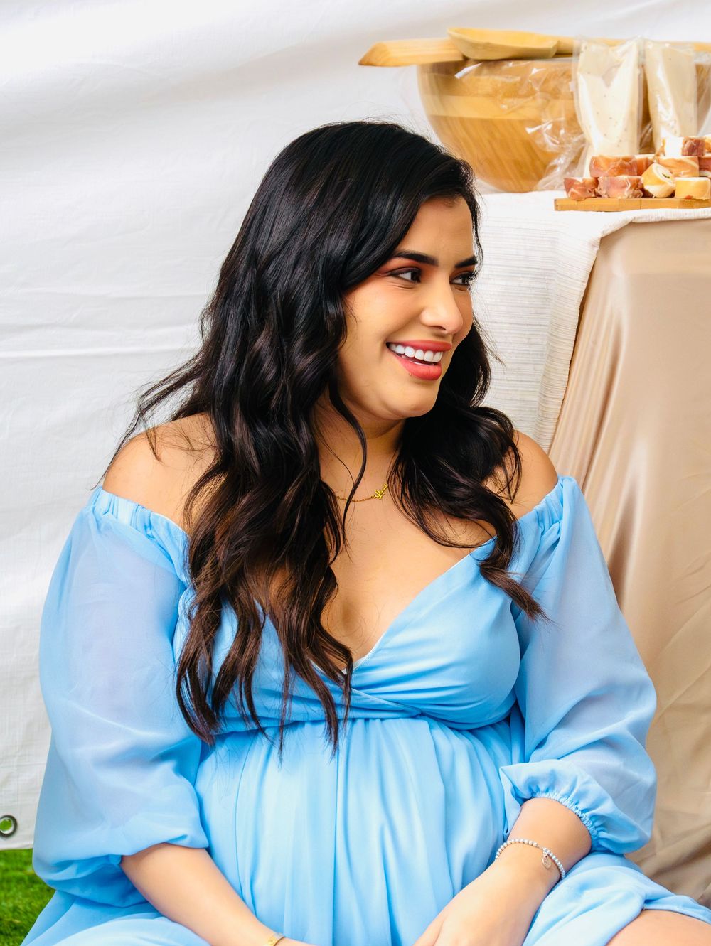 Smiling pregnant woman in a light blue maternity dress sitting at an outdoor baby shower celebration.