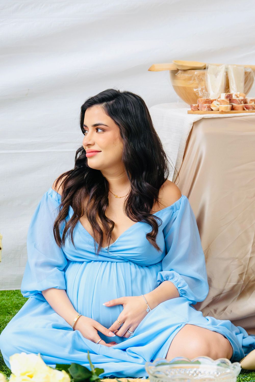 Pregnant woman in blue off-shoulder dress sitting gracefully at a baby shower, smiling and cradling her baby bump.