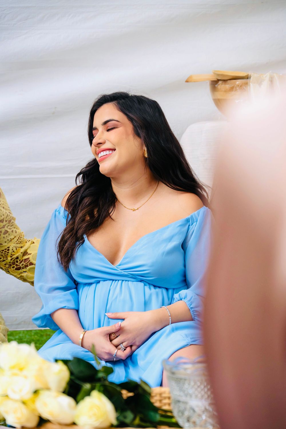 Joyful pregnant woman in a blue off-shoulder dress laughing at her baby shower with white roses nearby.