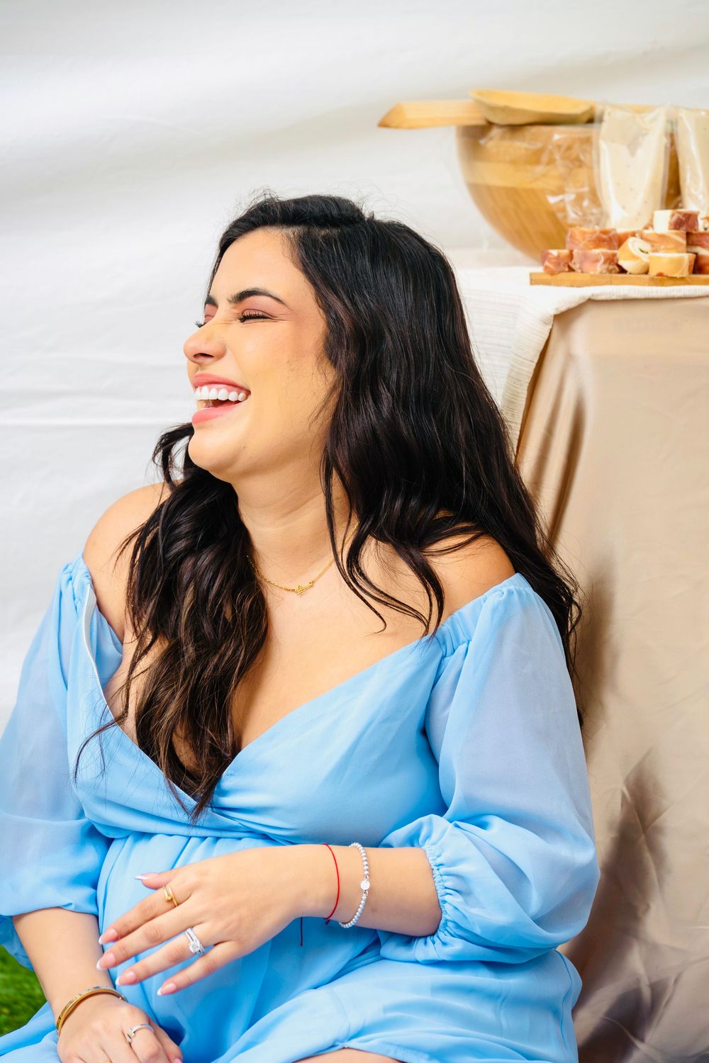 Laughing pregnant woman in a blue maternity dress sitting at a beautifully styled baby shower picnic.