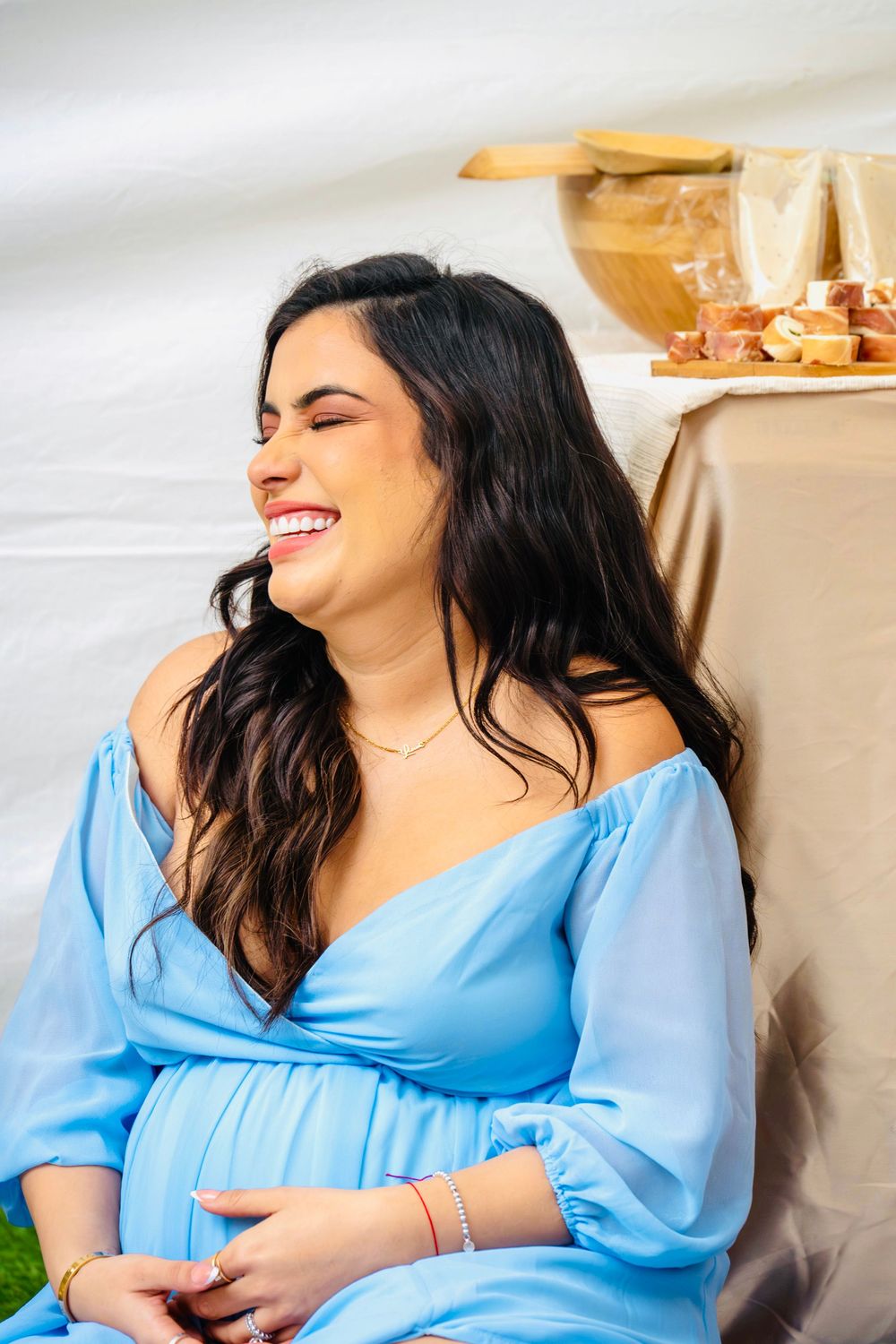 Radiant pregnant woman in a light blue off-shoulder dress laughing joyfully at her outdoor baby shower.