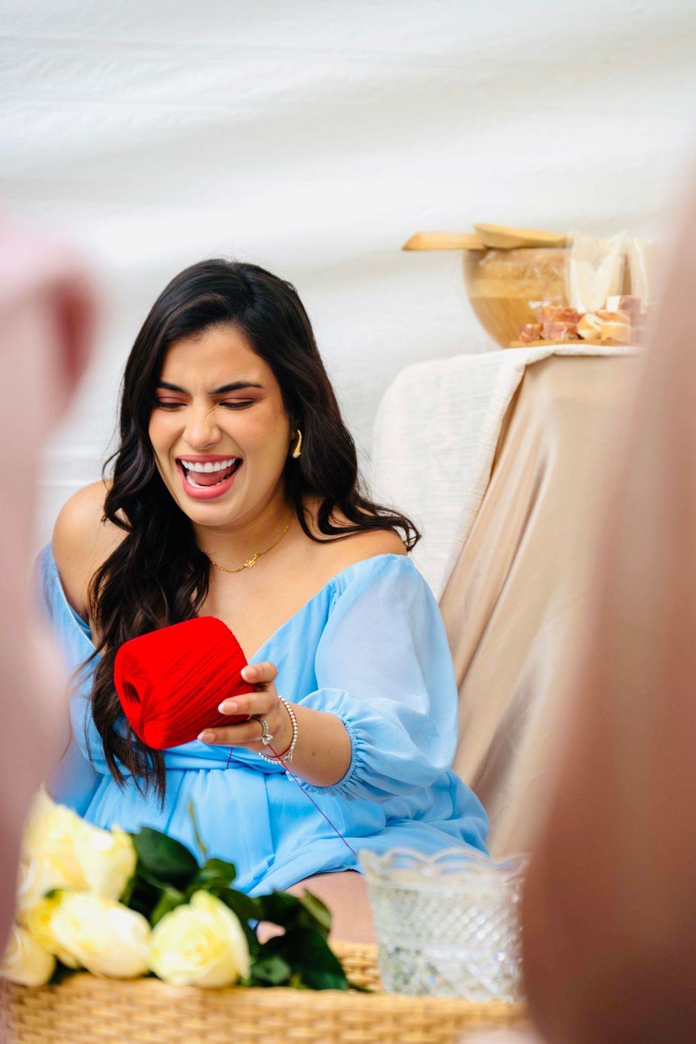 Excited woman in blue off-shoulder dress laughing while opening a red ring box at a celebration.