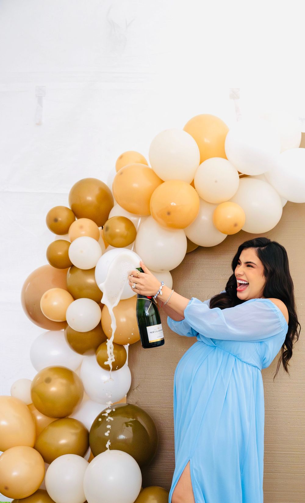 Pregnant woman in blue dress joyfully popping champagne bottle in front of gold and cream balloon arch.