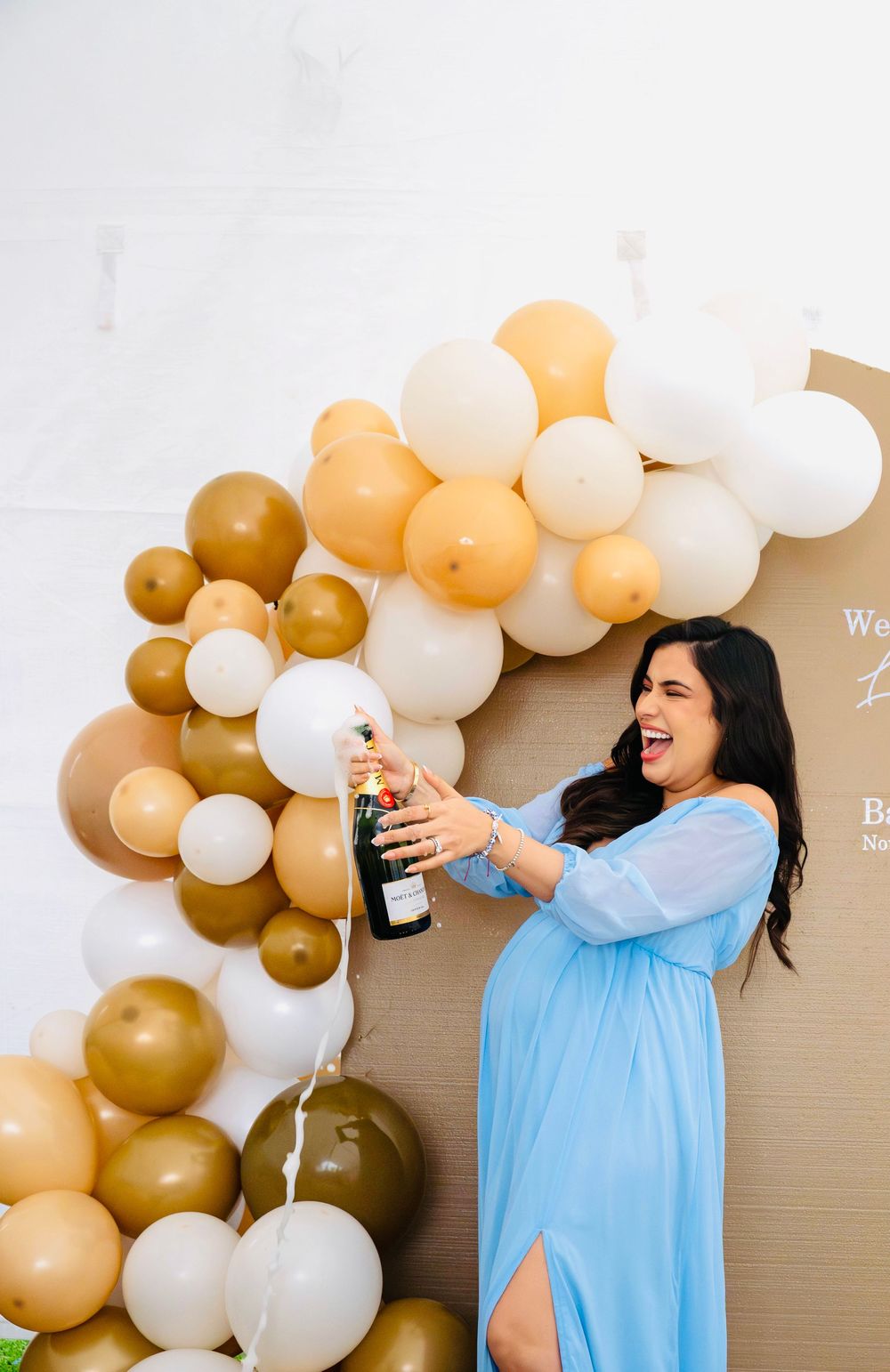 Laughing pregnant woman in blue dress popping champagne bottle at baby shower balloon backdrop.