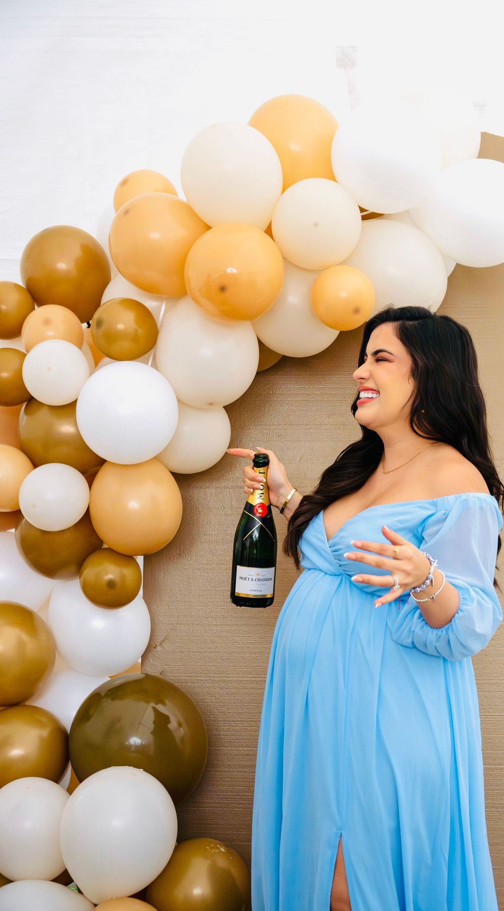 Smiling pregnant woman in blue maternity dress holding Moet champagne at gold balloon display.