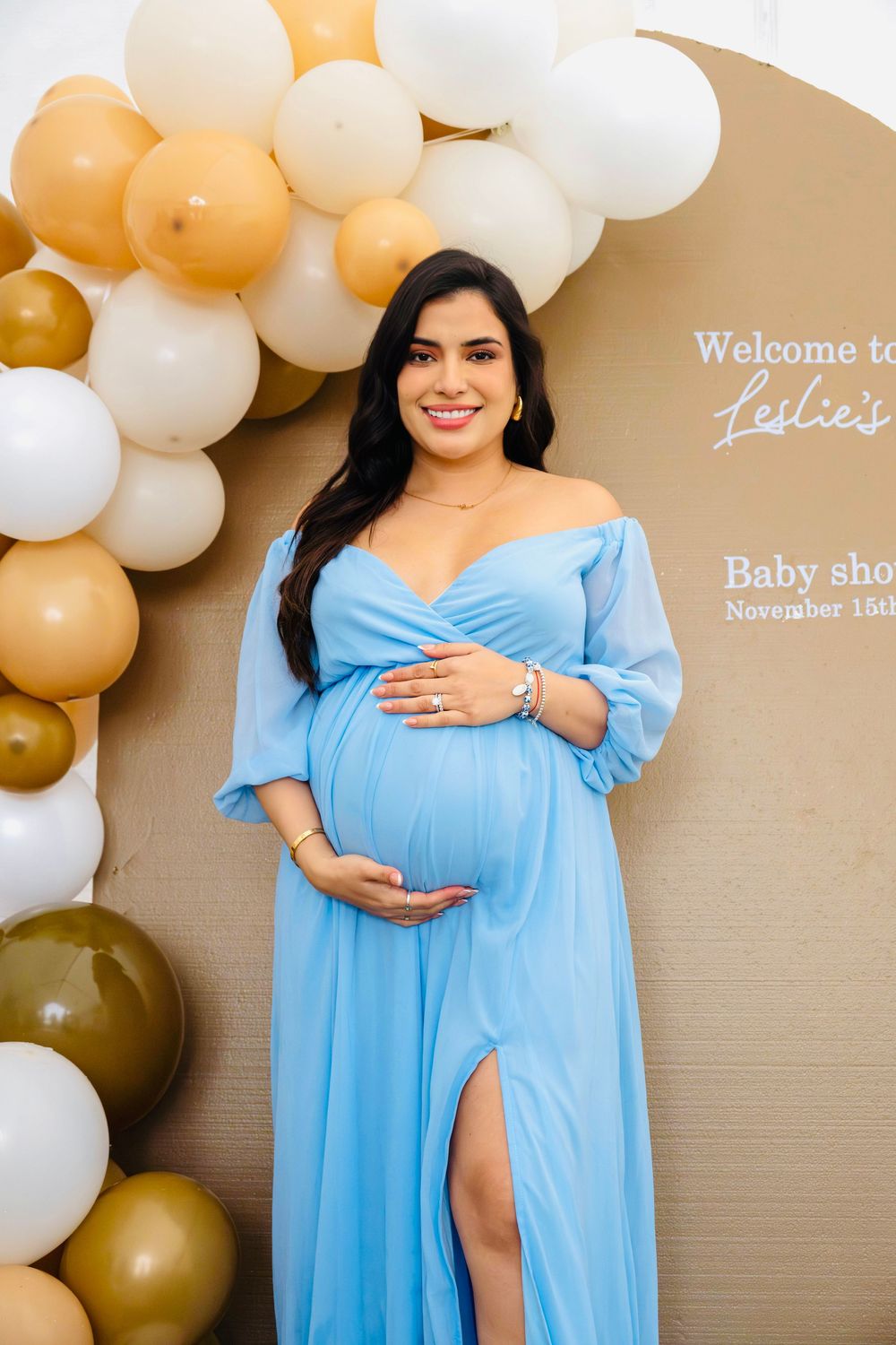 Pregnant woman in blue off-shoulder gown cradling baby bump at Leslie's baby shower backdrop.