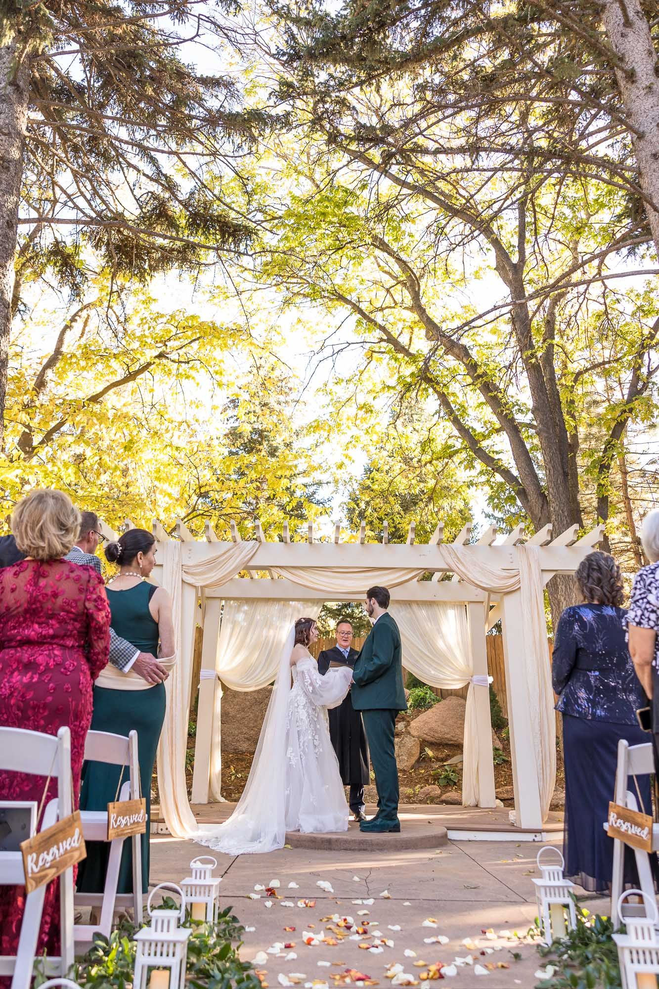 A wedding ceremony takes place at a charming Victorian-style venue with autumn trees and rose petals scattered along the path.
