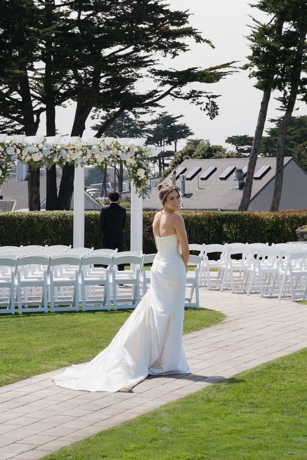 A bride in a white wedding dress walks along a stone path with white chairs set up for an outdoor ceremony.