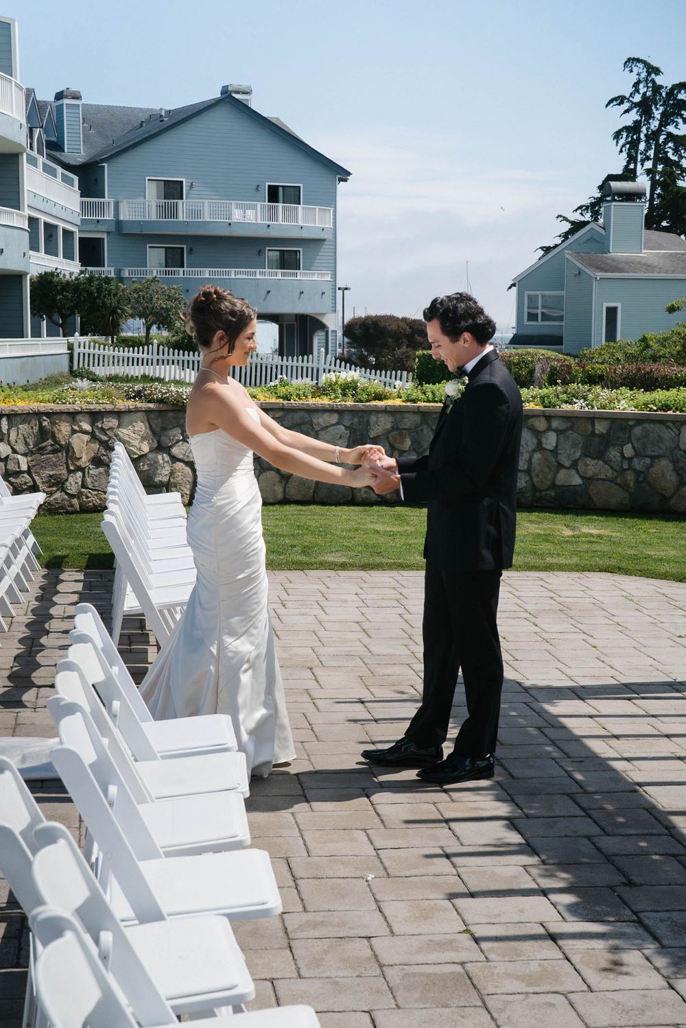 Couple dances together on an outdoor stone patio with white chairs and coastal homes in the background.