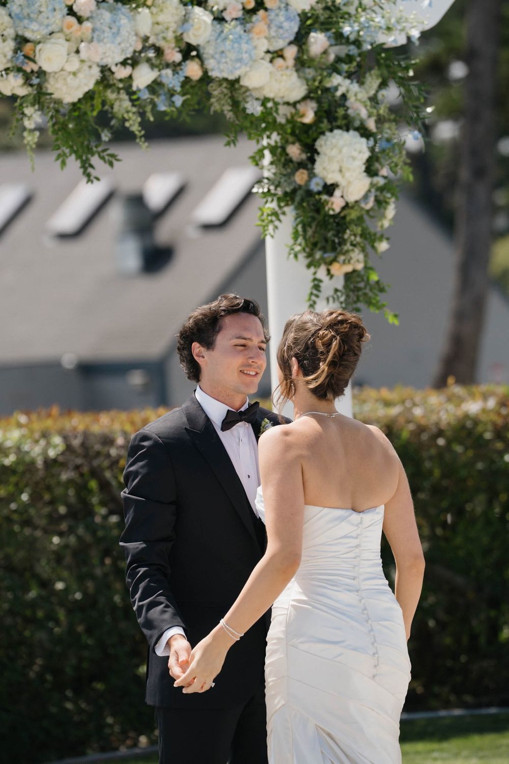 White floral arrangements frame an outdoor wedding scene with a stone patio and coastal backdrop.