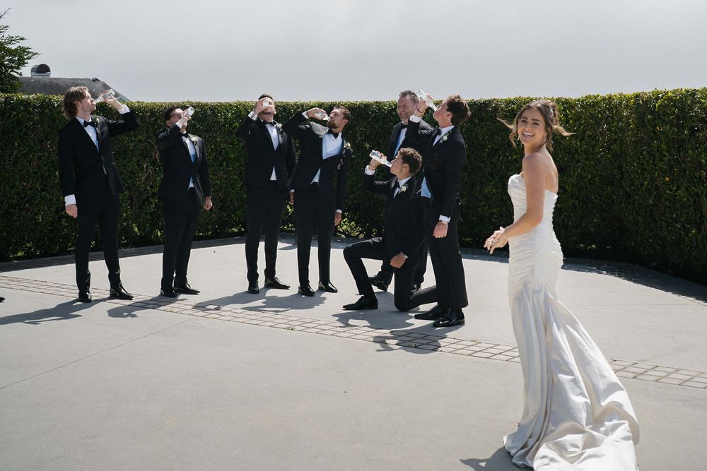 A group of people in formal attire stand in line outdoors on a sunny day for a wedding ceremony.