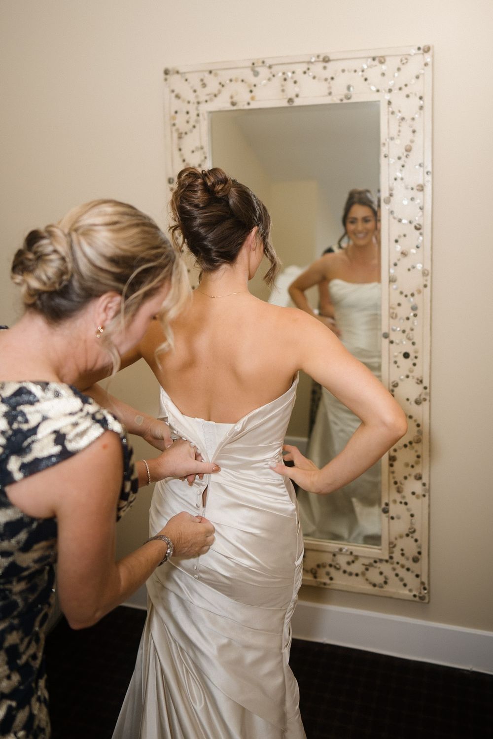 A person helps adjust the back of an elegant white wedding dress as the wearer looks in a decorative mirror.