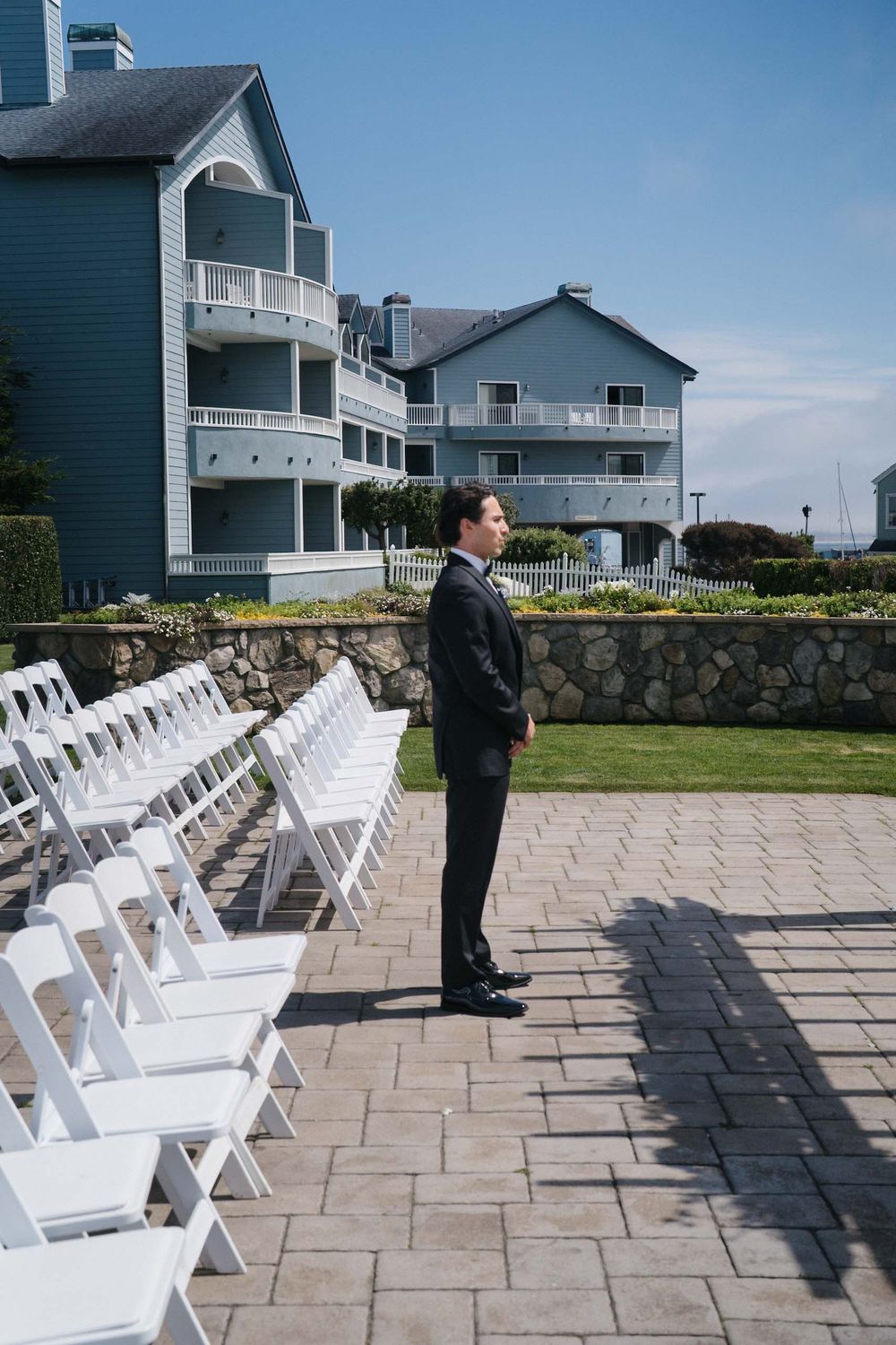 White chairs arranged in rows on a stone patio for an outdoor ceremony with coastal homes in the background.