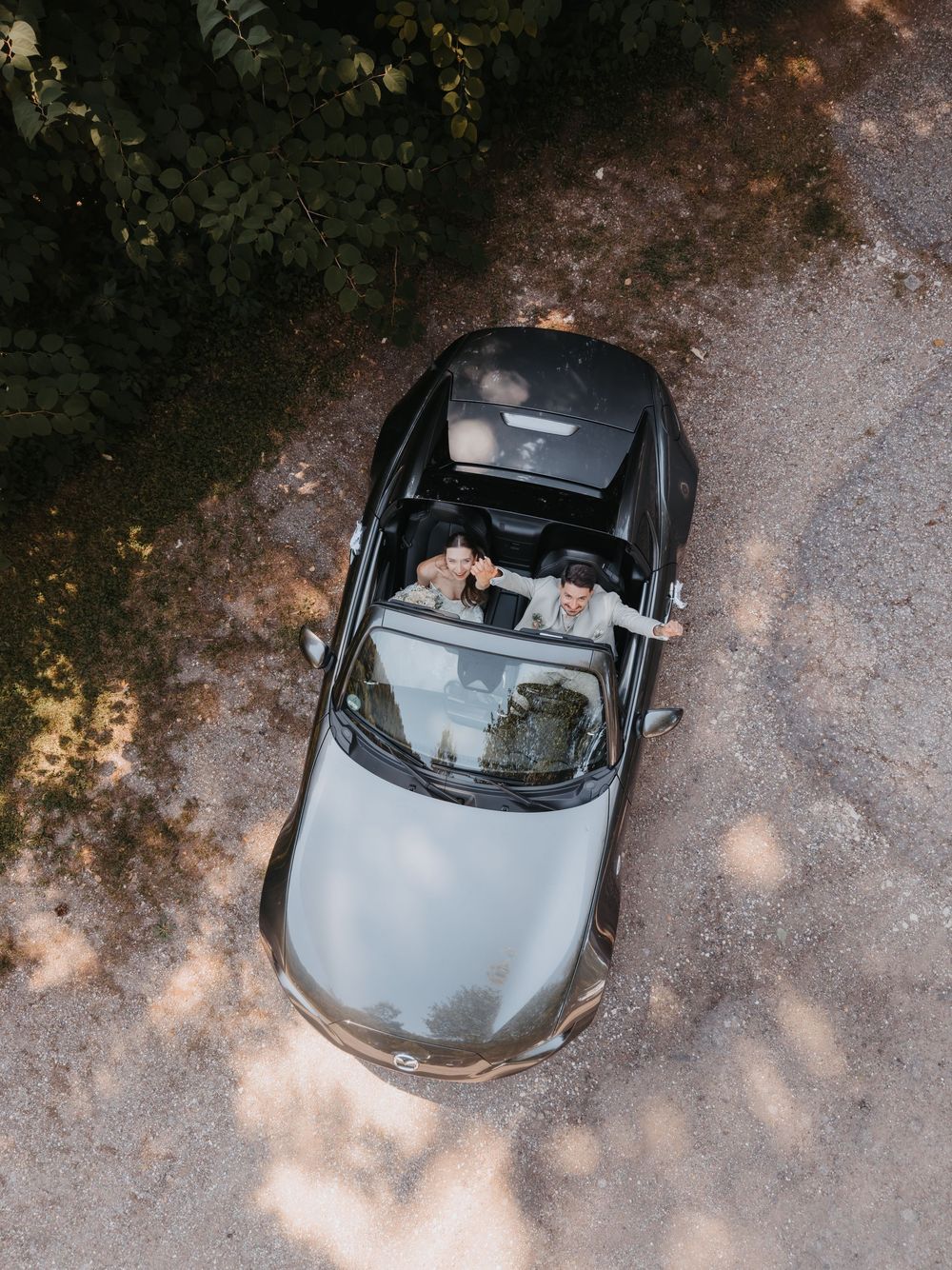 Aerial view of two people in a black convertible car on a gravel road surrounded by trees.