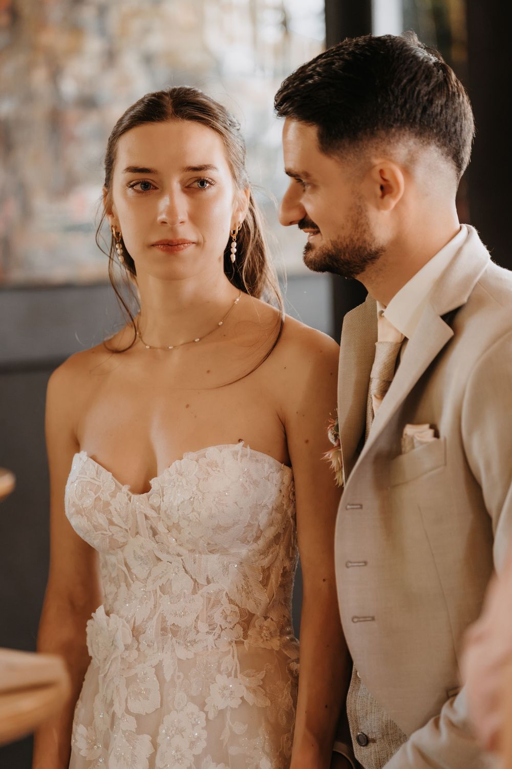 Bride in floral strapless gown stands beside groom in beige suit, sharing an intimate moment at their wedding.