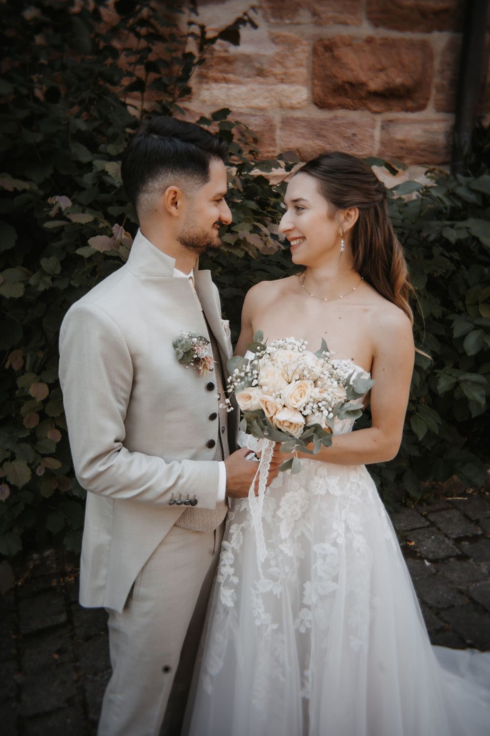 Bride and groom in elegant white wedding attire share a romantic gaze, holding a bouquet of roses outdoors.