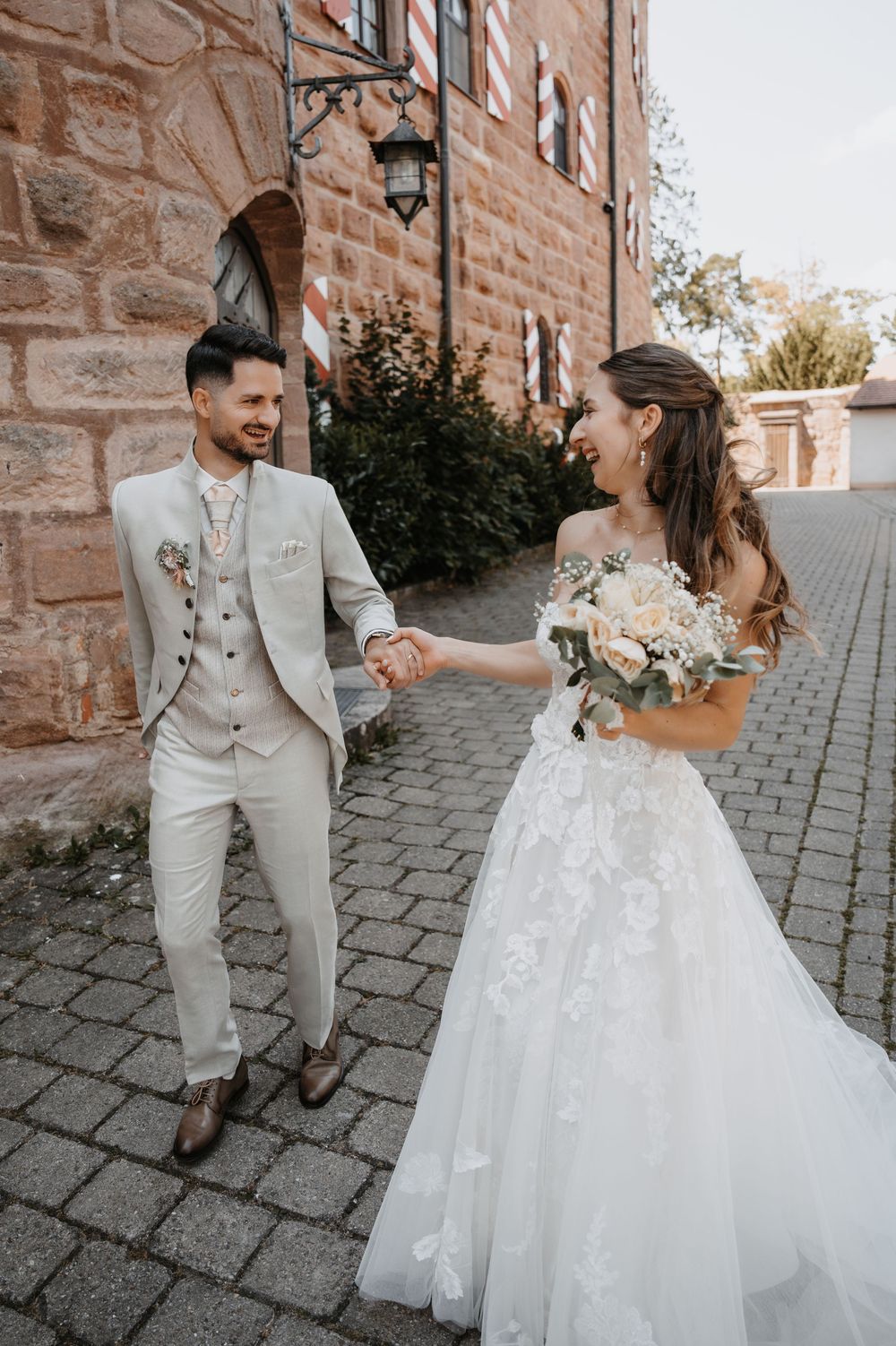 Bride and groom holding hands smiling at each other outdoors near a stone building, bride in lace gown holding bouquet.