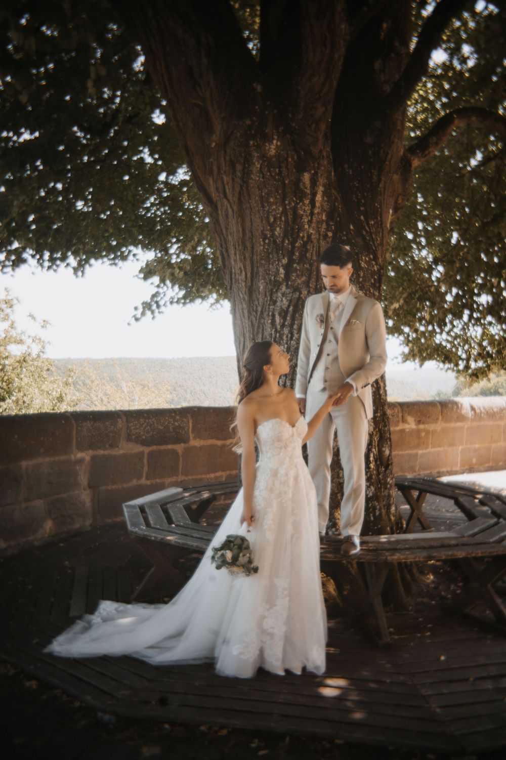 Bride and groom holding hands under a large tree, bride in lace gown, groom in tan suit, scenic outdoor backdrop.