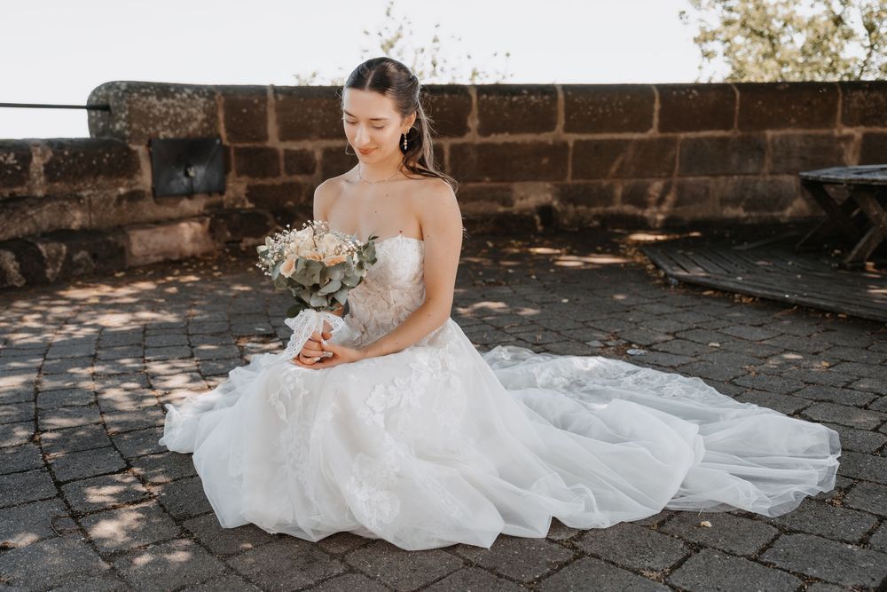 Bride in a strapless ball gown wedding dress holding a bouquet, seated on stone pavers outdoors.