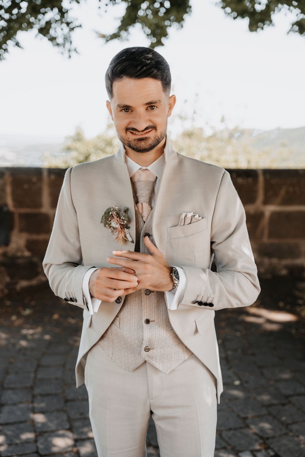 Smiling groom in beige suit holding boutonniere outdoors on sunny wedding day.