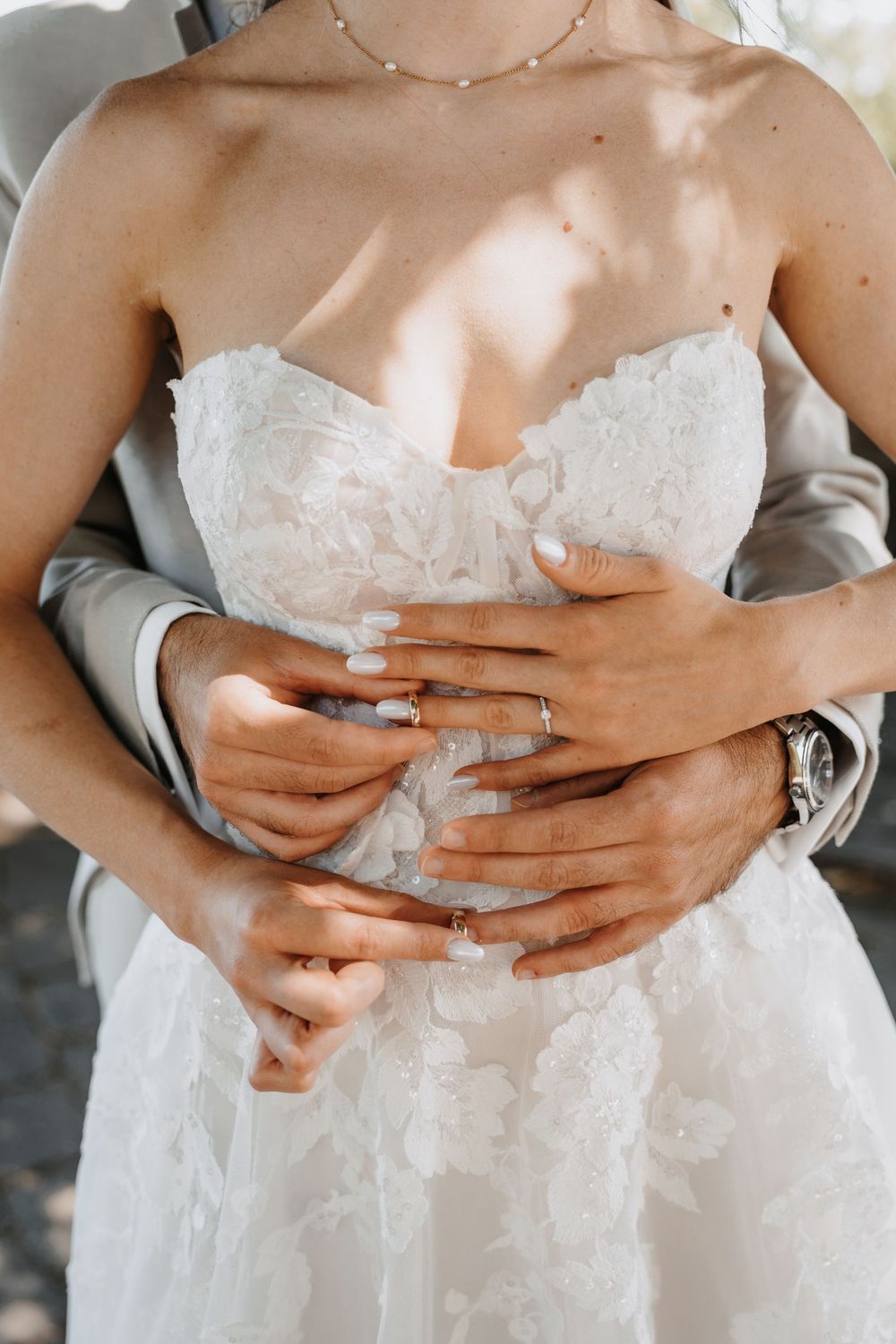 Close-up of groom embracing bride from behind, both wearing wedding rings, bride in strapless lace wedding dress.