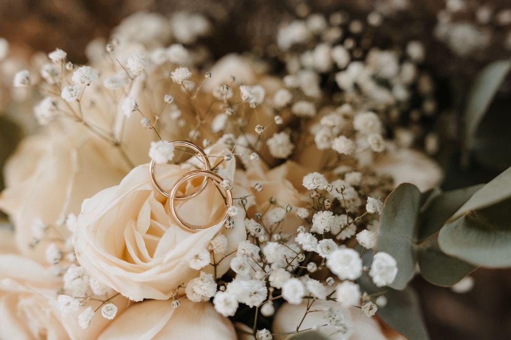 Two gold wedding rings rest on a bridal bouquet of cream roses and white baby's breath flowers.