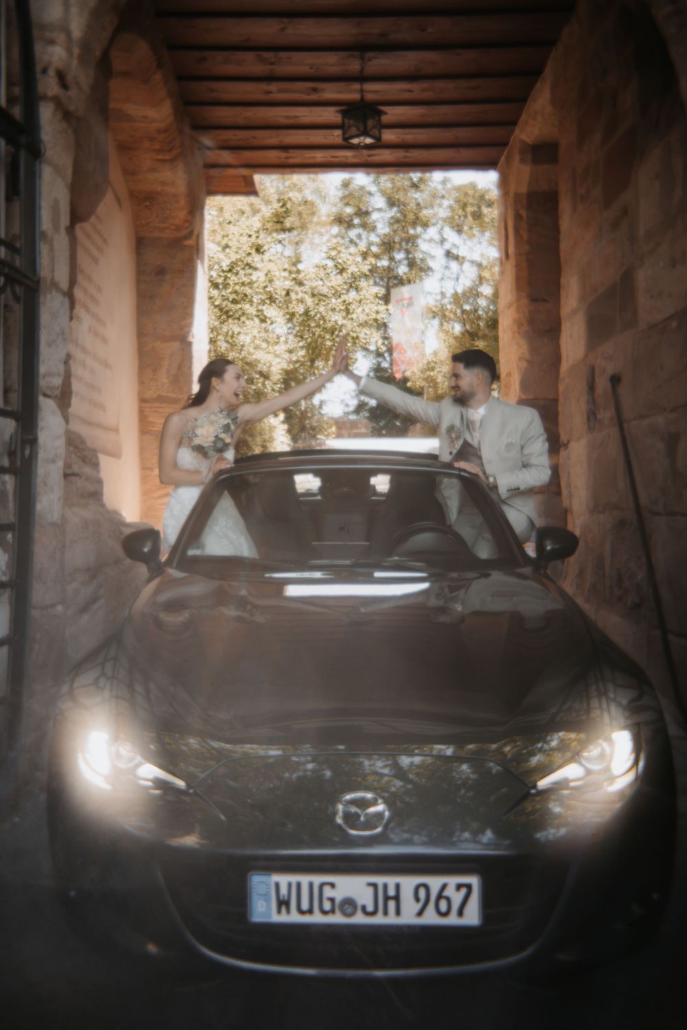Bride and groom standing behind a black Mazda convertible in a stone archway on their wedding day.