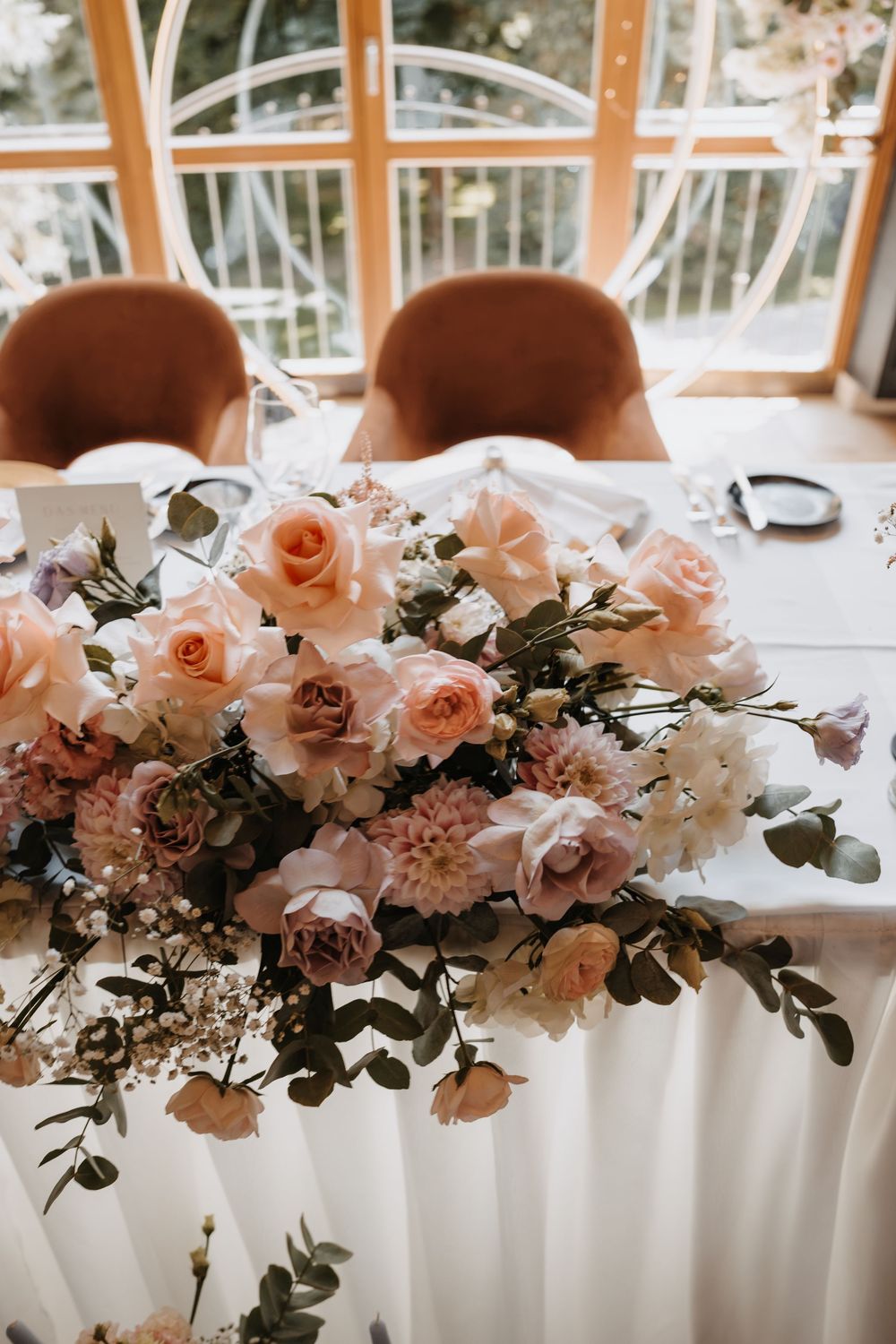 Elegant wedding table centerpiece with blush pink roses, dahlias, and greenery arrangements in a bright venue.
