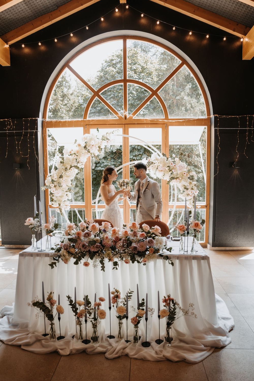 Bride and groom toasting at floral sweetheart table with white draping and pink blooms before arched window.