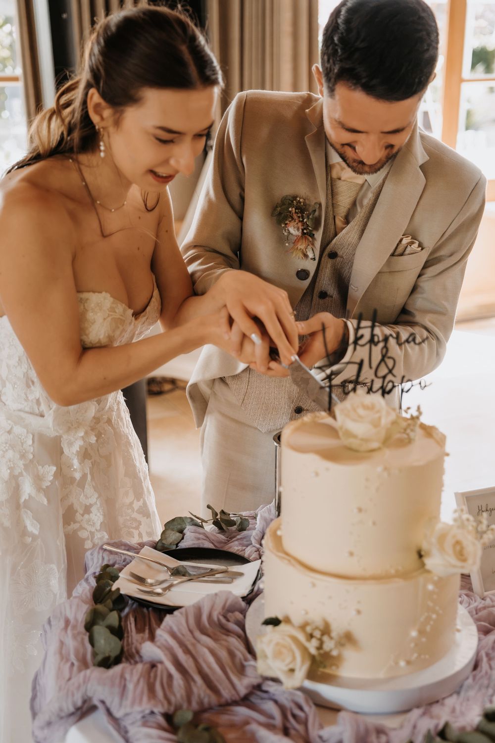Bride and groom cutting elegant two-tier white wedding cake decorated with roses.