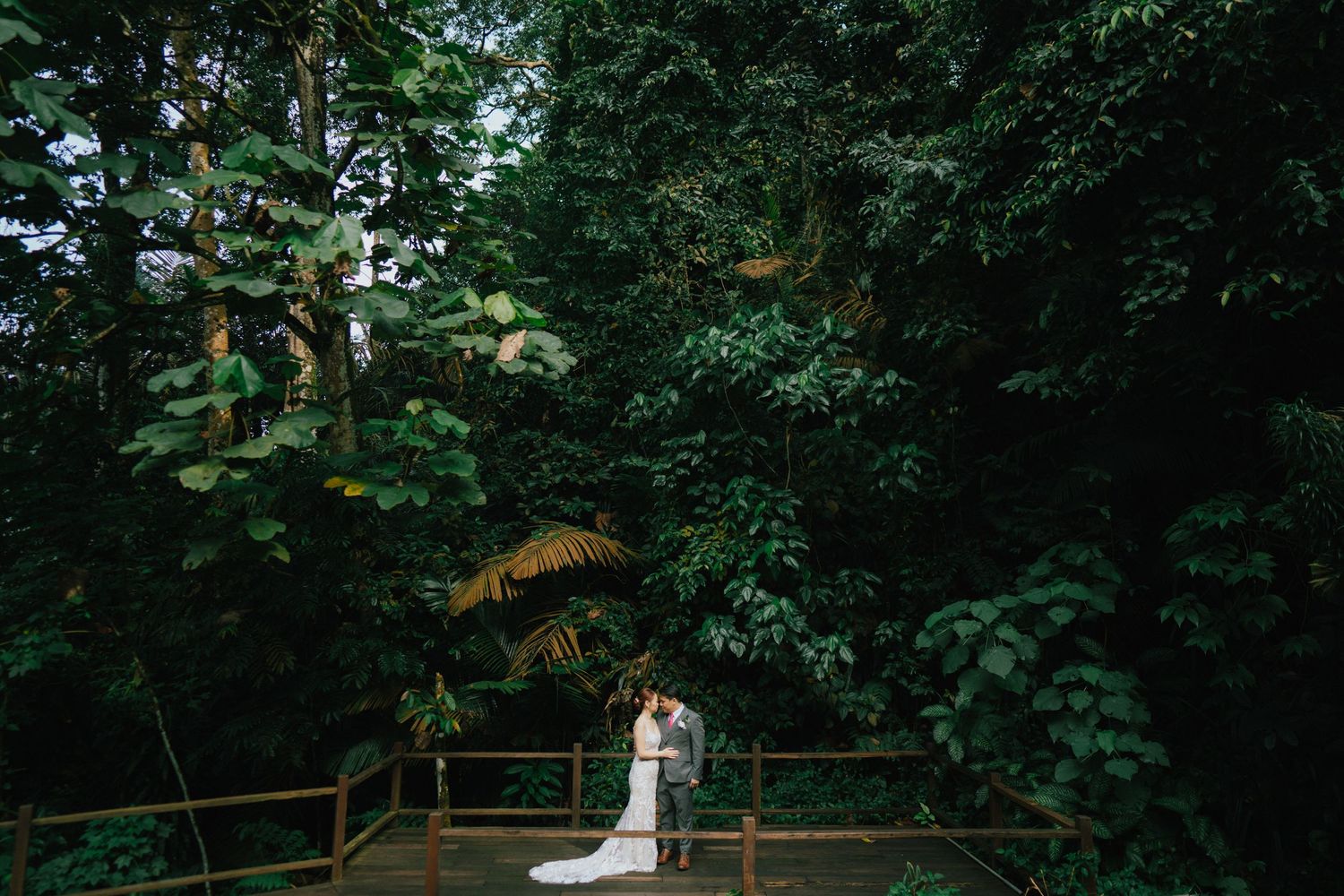 Bride and groom share a romantic embrace on a wooden deck surrounded by lush tropical forest greenery.