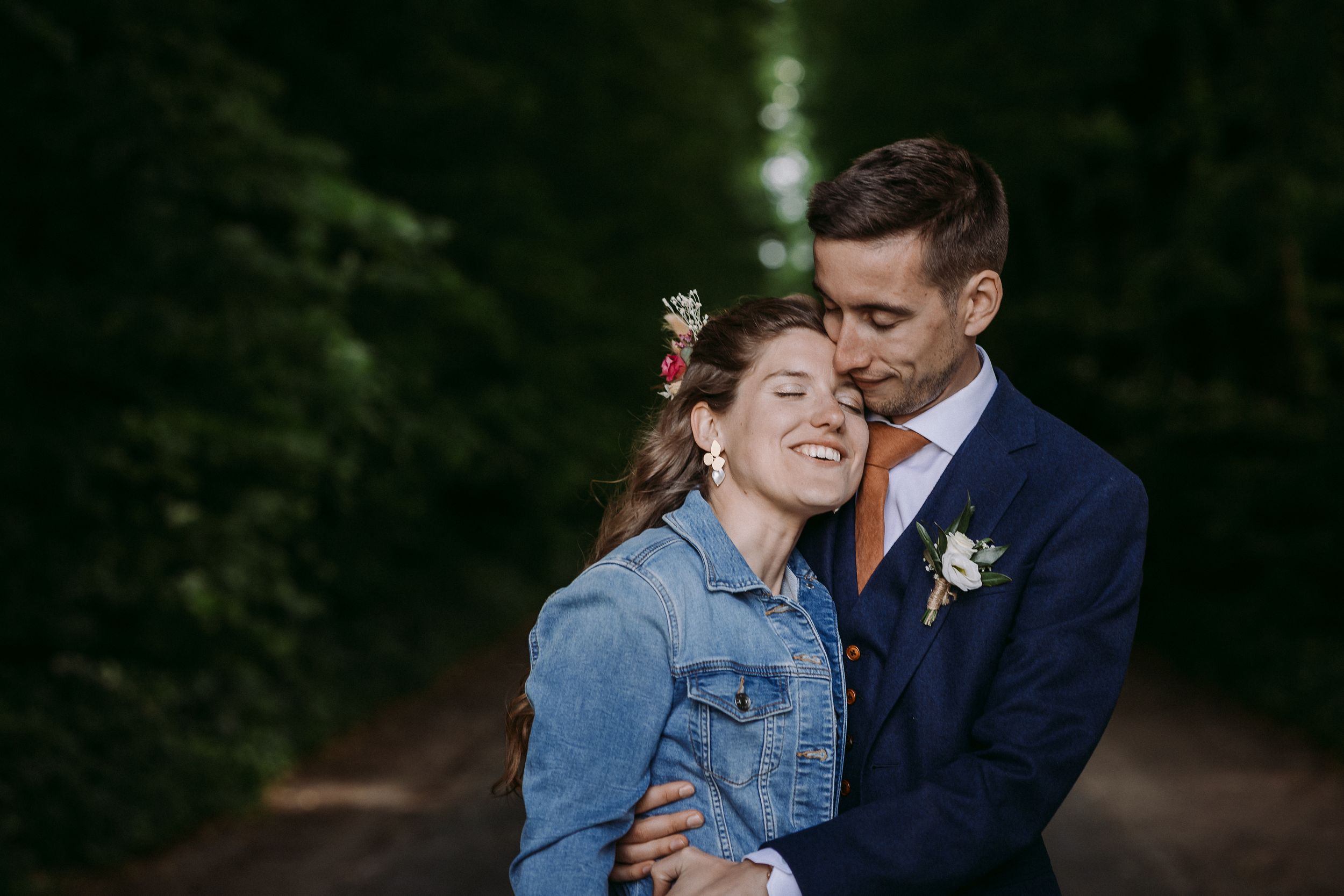 A couple embraces on a forest path, with the bride wearing a denim jacket over her dress and groom in navy suit.