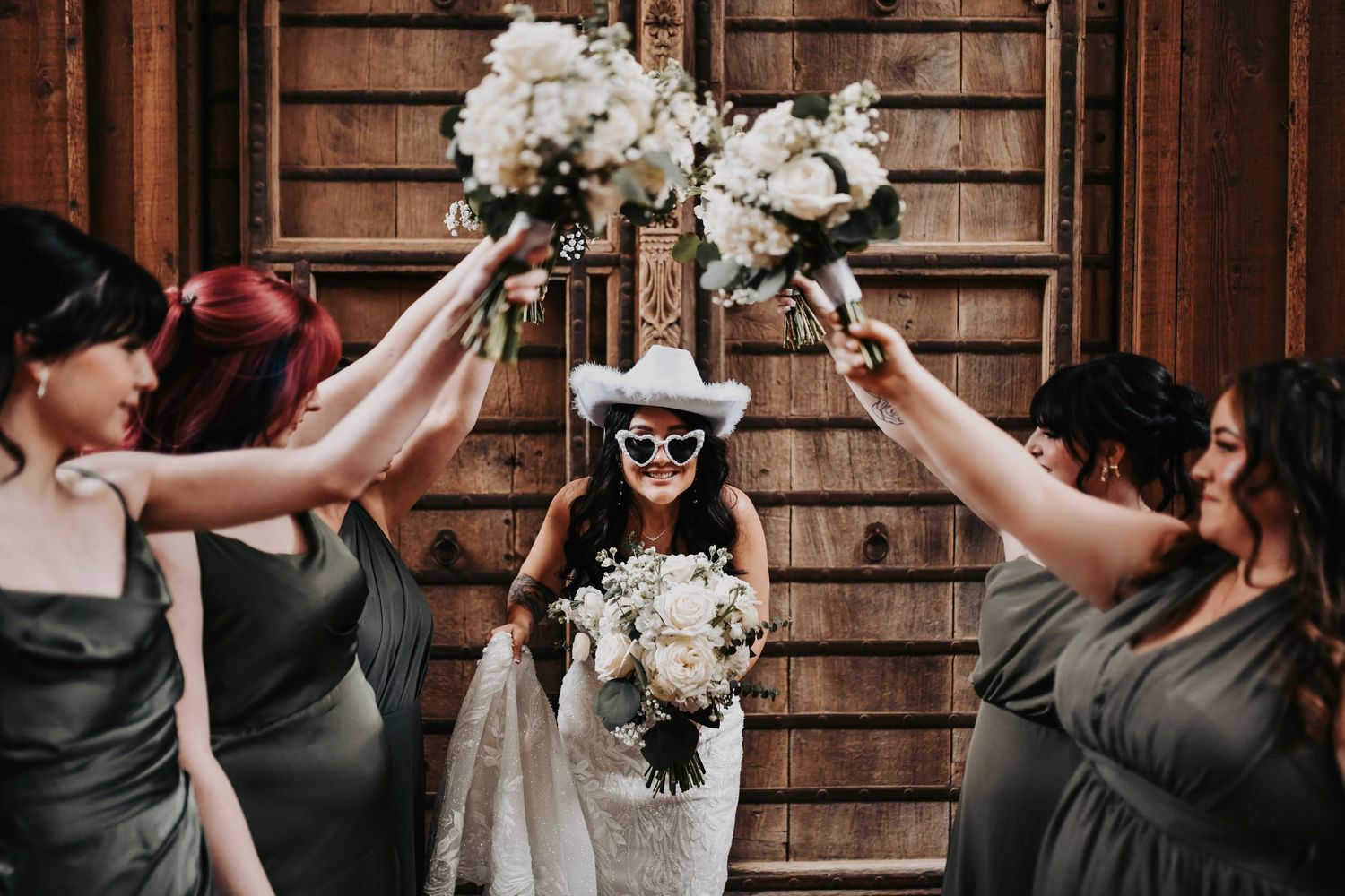 Candid photo of bridesmaids in olive gowns laughing with the bride while holding organic greenery bouquets.