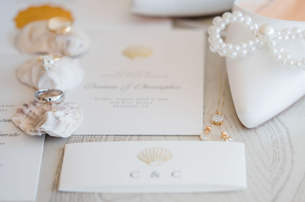 Elegant wedding details including white bridal shoes, pearl jewelry, and delicate invitations arranged on light fabric.