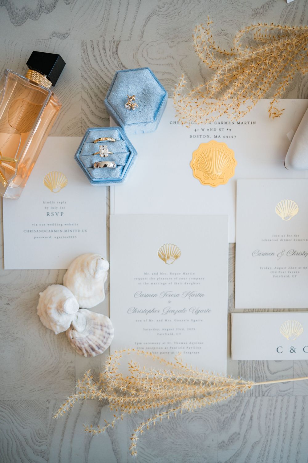 Wedding invitation suite with seashell motifs in soft blue and gold tones displayed with dried flowers and pearls.