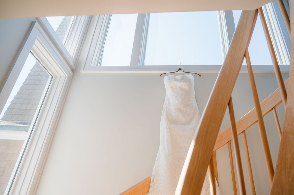 A white wedding dress hangs from a hanger near tall windows and wooden staircase railings in natural light.